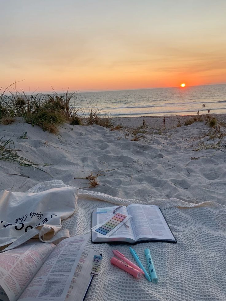 A peaceful beach scene at sunset with a view of the ocean, featuring a Bible open to a page, colorful highlighters, and a color swatch on a blanket with grass in the foreground.