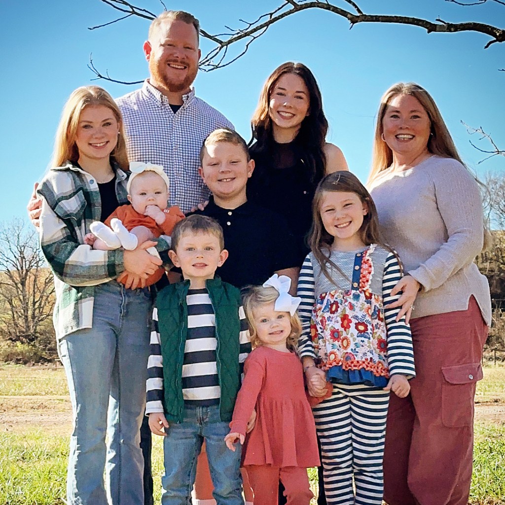 A smiling family posing for a picture outdoors, featuring a mix of adults and children. The backdrop includes trees and a clear blue sky.