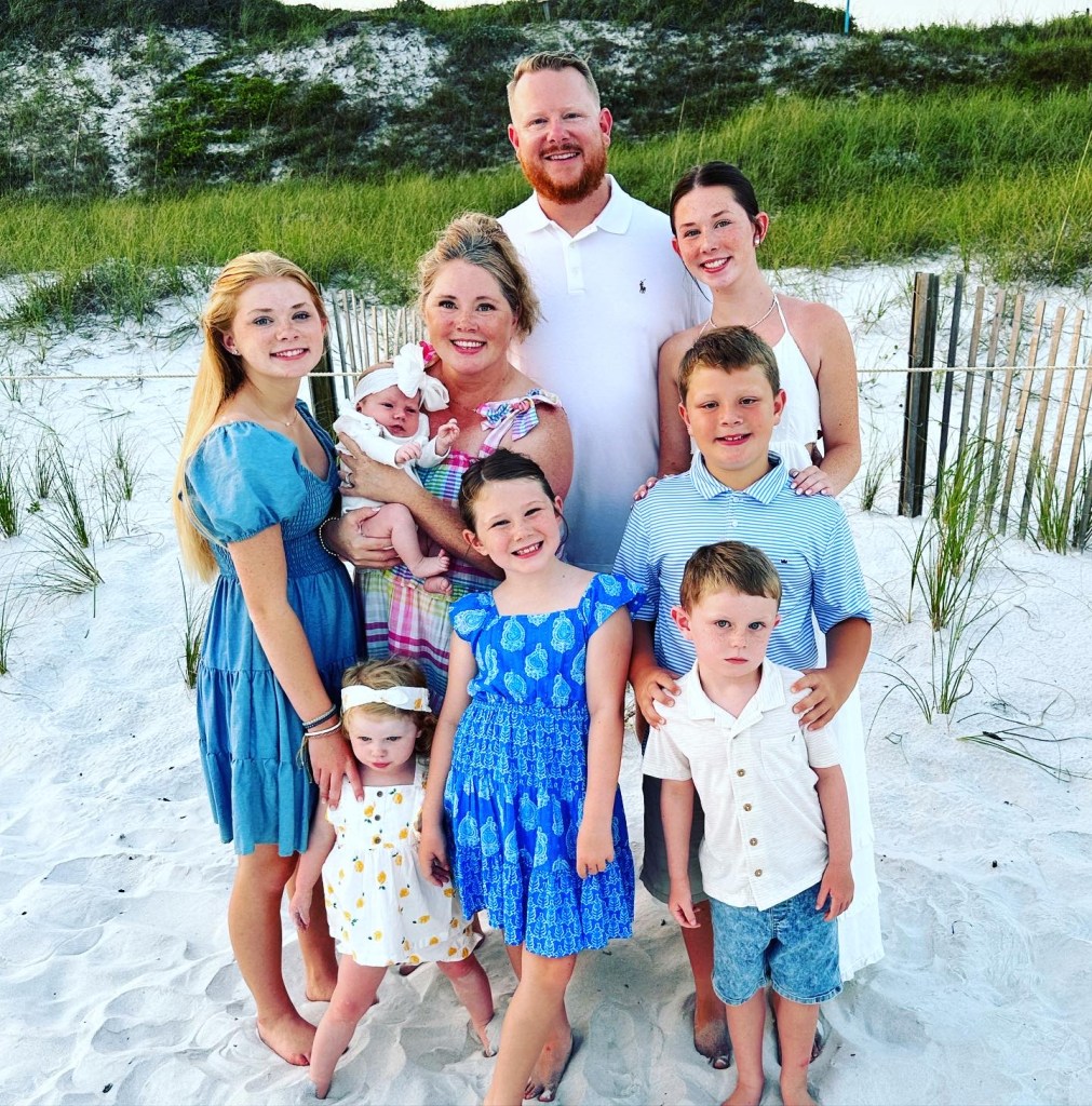 A family of eight poses together on a beach, with green grass in the background. The group includes adults and children, all wearing summer outfits. One adult is holding a baby.
