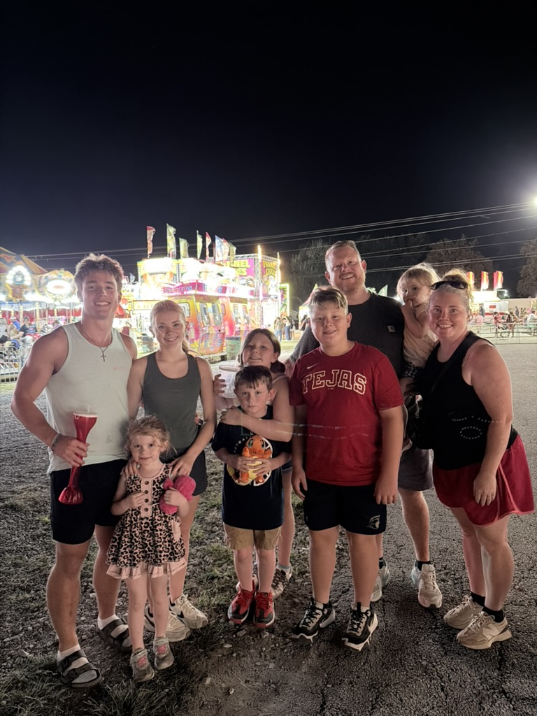A group of seven people, including adults and children, posing together at a nighttime fair or carnival, with colorful rides and lights in the background.