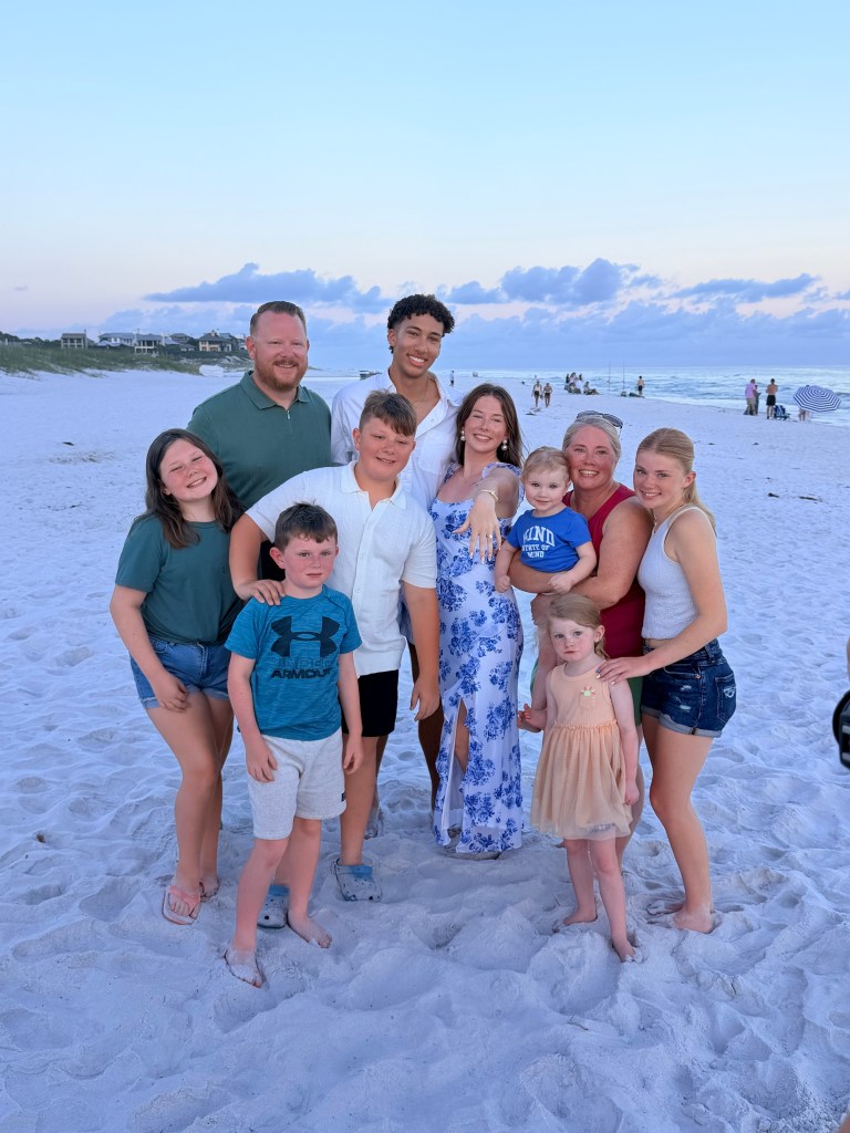 A group of nine people of various ages poses together on a beach at sunset, surrounded by soft white sand, with the ocean and clouds in the background.