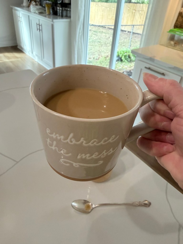 A person holding a large coffee mug that says 'embrace the mess' with a spoon placed on a table in the foreground, and a window with a view of a backyard in the background.