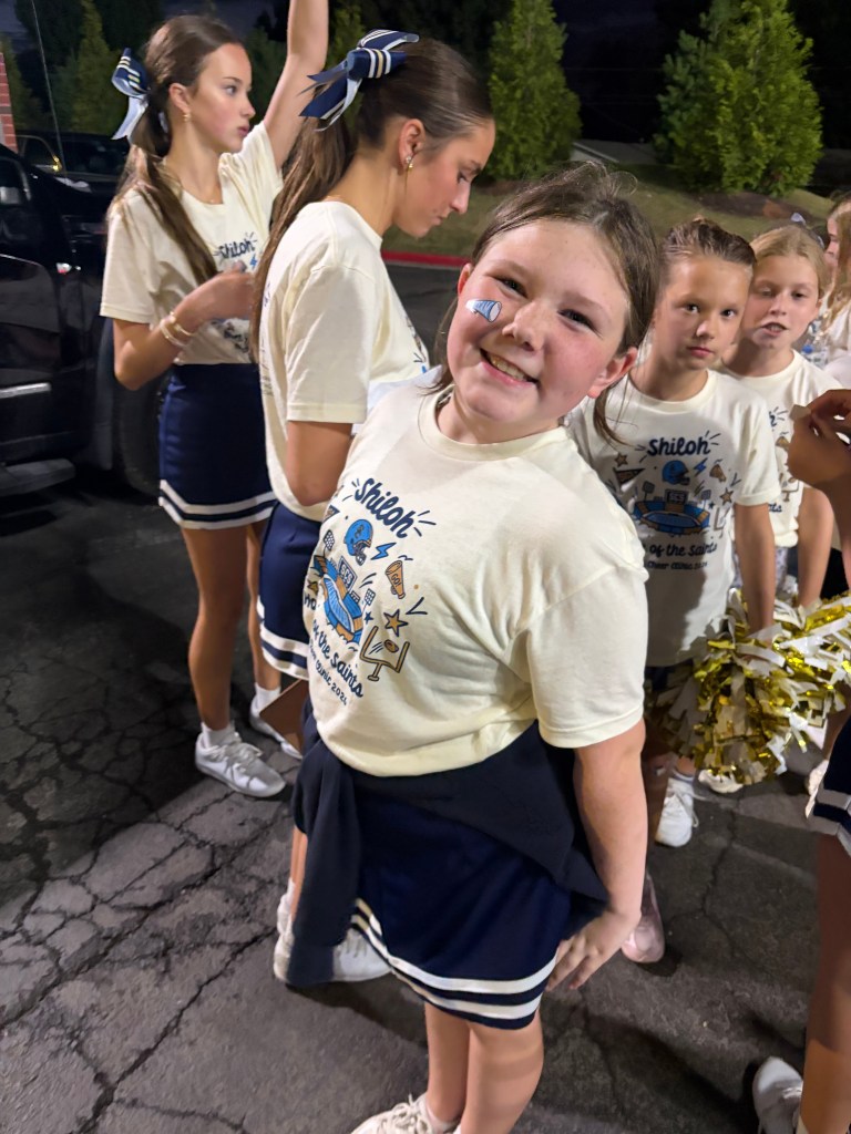 A group of young cheerleaders in matching outfits, with one girl in the foreground smiling and showing face paint while others prepare in the background.