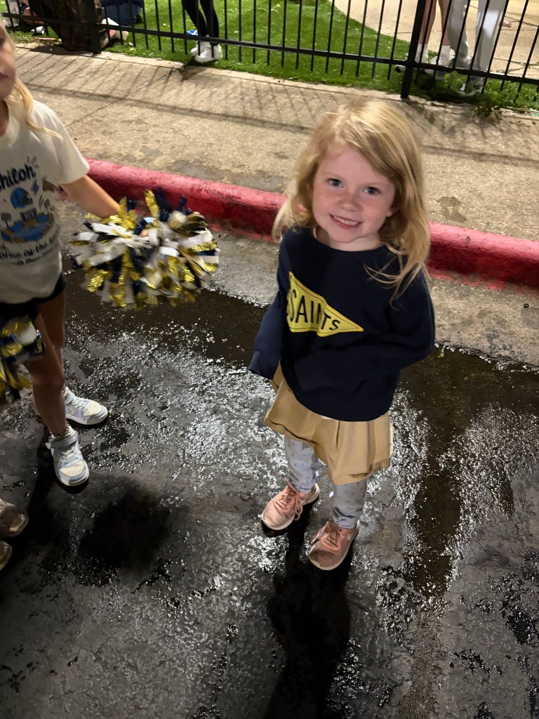 A young girl with wavy blonde hair smiles while standing on a wet surface, wearing a dark sweater with 'SAINTS' printed on it and a beige skirt, holding a cheerleading pom-pom. Other children and spectators can be seen in the background.