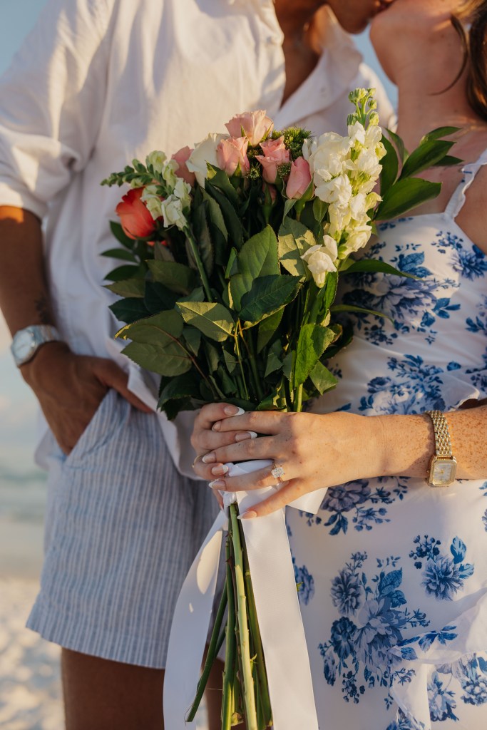 A close-up of a couple embracing, with a bouquet of pink and white flowers held by the woman, wearing a floral dress, while the man stands beside her in casual attire.