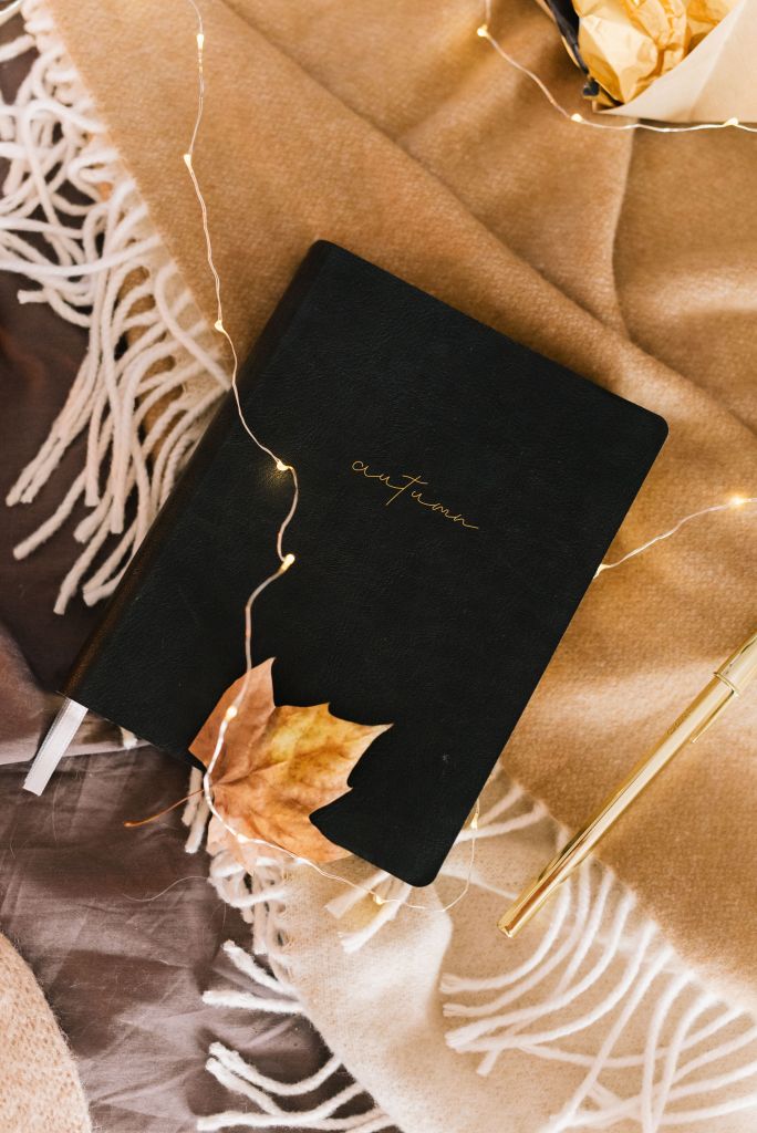 A black journal with the word 'autumn' embossed in gold, placed on a cozy beige blanket with delicate white fringes. A dried leaf and a gold pen are nearby, along with fairy lights adding a warm glow.
